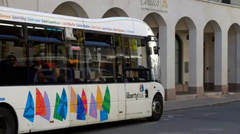 BBC A LibertyBus in Jersey. It is parked on a city street and is white with a series of colourful abstract sail-like shapes painted along its lower side. It also has large windows showing passengers inside. Above the windows, various place names are listed.