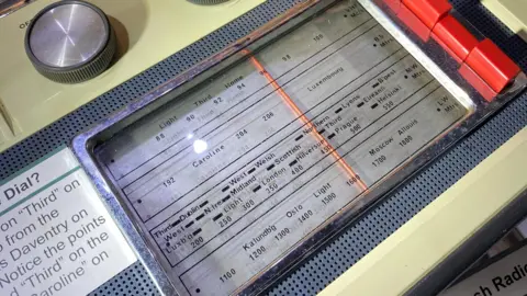 Daventry Museum Old cream-coloured radio with frequency window revealing a red needle which can be moved across a screen showing frequencies and locations. There is a black and silver rotating knob above the screen and an information sticker to the left.