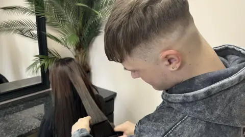BBC/Louise Hobson A woman with long brown hair sits in a chair facing a mirror with her back to the camera on the left. On the right is Ashton, a young man wearing a grey hoodie, with short brown hair. He is straightening the hair of the woman 