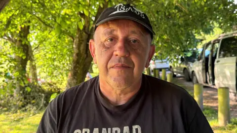 Taylor's grandad Andy stood outside under a tree. He is looking straight at the camera and is wearing a black t-shirt and hat.