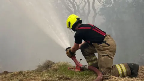 A firefighter wearing a fluorescent yellow helmet and uniform directs a jet of water from a red hose. He kneels on a grassy mound with trees in the background and is surrounded by hazy smoke.