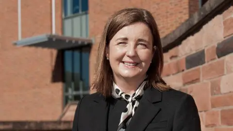 Shropshire Council A woman with long brown hair, a black jacket and white scarf with black spots in front of a red brick wall