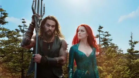 Jason Momoa as Aquaman looks towards us, holding a five-pronged trident/spear in his hands. He wears a dark vest and has several tattoos on his arm. To his left, a woman with long red hair, wearing a green dress, looks at him. Behind them are several trees.