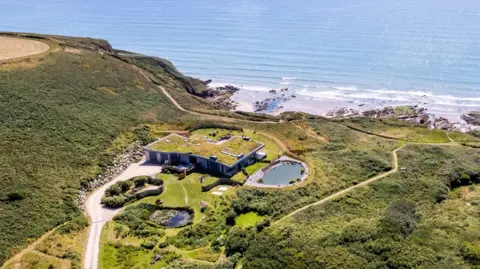 Fine & Country Aerial view of a modern, single-story house with a grass-covered roof nestled into a coastal hillside.