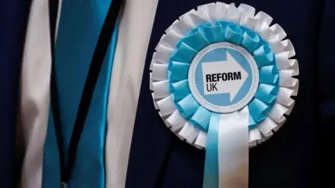 Close-up photo of a blue and white Reform UK rosette worn by someone wearing a dark suit. 