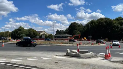 Sofia Luis-Hobbs/BBC Vehicles pass through a partially reconstructed roundabout, with traffic cones and temporary barriers marking ongoing roadworks. Excavators and construction equipment are working in the central island area, while unfinished kerbs and paving are visible in the foreground. Trees and low buildings line the background beneath a bright blue sky with scattered clouds.