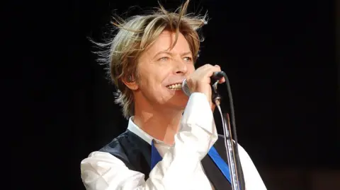 Getty Images David Bowie singing into a microphone on stage. He has blonde hair and is wearing a black waistcoat and white shirt