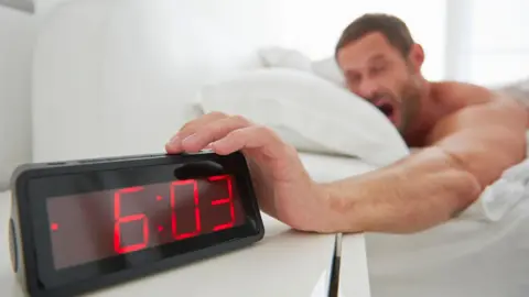 Getty Images A man lying in bed yawns as he reaches for a clock radio that shows the time 6:03 in red digits.