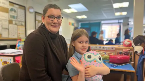 Woman with brown hair and brown jumper sat in a classroom next to her child, a little girl with a stuffed toy waving at the camera