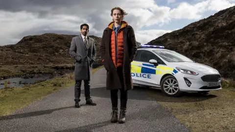 Black Camel Pictures/BBC Alba/John Maher Radia, dressed in long grey coat, and Groundsell, wearing a woollen coat over an orange jacket, are standing in front of a police car on a single-track island road. 