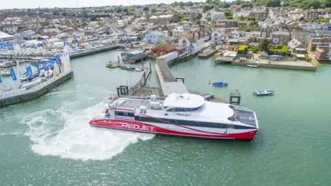 Red and white ferry at pontoon with buildings around the ferry terminal.