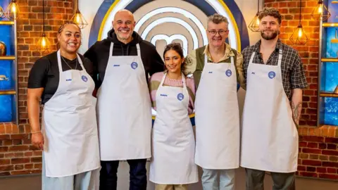 Five people - two women and three men - all wearing white aprons, stand in front of a brick wall.