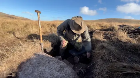 A man is leaning over in a trench sounded by a large boulder and very dry grass. There is a spade to one side.