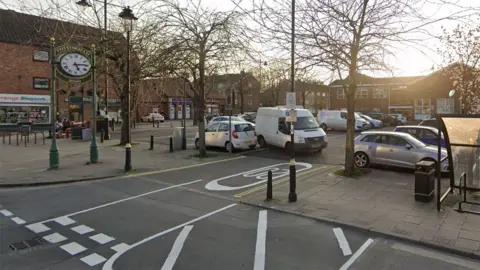 Google Google street view of Cottingham's Market Place car park with a large green clock with the name Cottingham on it and a number of vehicles parked in bays in a smart-looking square.