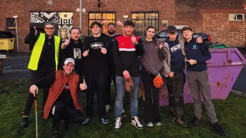 A group of eight men and one woman smiling at the camera with their thumbs up. They are stood in front of a purple skip