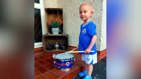 Claire Reaney A small blonde boy stands in a blue football kit with a tiny drum in front of him. He holds drum sticks in his hands and stares off camera.