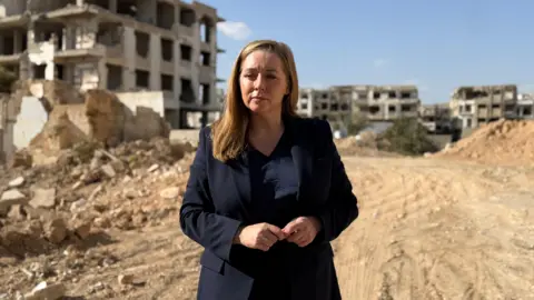 A blonde haired woman in a black suit stands in the middle of a dusty roadway. Her hands are clasped as she looks to the side of the camera. Behind her are three large, crumbling buildings and various piles of rubble. There is a blue sky above the damaged buildings.