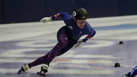 A man in a full lycra skating suit which is blue with union jack flags on. He is wearing a black helmet and sunglasses.