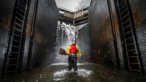 A volunteer wearing waterproof clothing stands in the drained canal with a pile of leaves on the end of a rake.