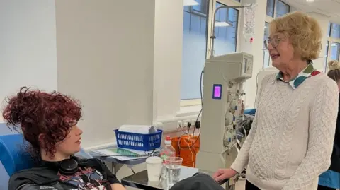 A young woman sat in a medical chair at a blood plasma donation centre looks up at an older woman who stands next to the bed. A blood collection machine is visible in the background.