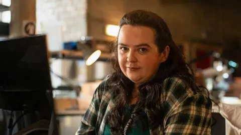 BBC Storrie in a scene from the programme Dinosaur looks directly at the camera. She is wearing a check shirt and a lanyard which says Glasgow. She is sitting in a deserted office with an anglepoise lamp seen behind her.