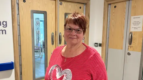 The image shows a person standing in a hallway outside a set of wooden double doors marked “Dining Room.” The person is wearing glasses and a red striped top with a heart design. Behind the doors are small window panels revealing part of a room inside, and there is a notice posted on one of the doors. The setting appears to be a care facility or hospital environment.