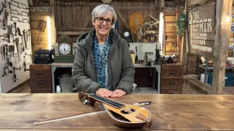A woman in a jacket and scarf sat behind a wooden table. A yellow lyra and its bow are resting on the table. She is sat on the set of The Repair Shop, with several antiques and tools visible in the background.