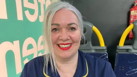 A woman in a navy nursing uniform sits on a bus seat. She is smiling. Her hair is blonde and tied back, with a loose strand by her face. She is wearing bright red lipstick.