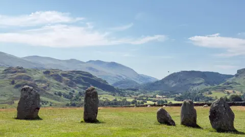 Jonny Gios Five standing stones - part of Castlerigg stone circle - on a grassy plain. Behind are mountains, green with trees and grass, under a hazy blue sky.