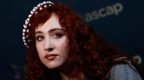 Getty Images Chappell Roan at a red carpet event. Chappell is a woman in her 20s with long, curly, dyed red hair and blue eyes. She wears a pearl-studded beret on her head and dark red lipstick. 