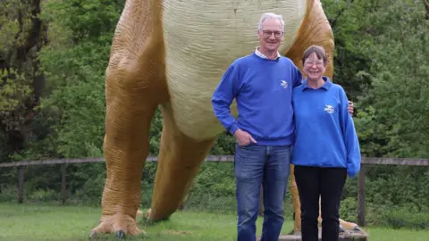Dinosaur Park Tenby Simon and Amanda Meyrick, both wearing glasses and blue jumpers, stand with their arms around each other in front of a large model dinosaur. 