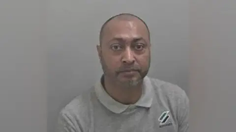 A man with a shaved head and stubble is wearing a grey polo shirt and looking at the camera in a police photograph.