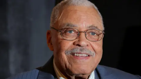 Getty Images James Earl Jones looks at the camera wearing round glasses and smiles. The actor has a gray mustache and short gray hair. 