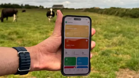 A close up picture of a man's hand, holding a mobile phone. An app can be seen opened on the phone screen. In the background, there are black and white cows standing in a green field.