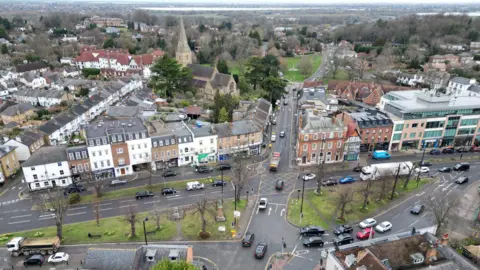 Aerial view of Esher town centre with a crossroads in the centre and in the distance a village green and church. The streets are lined with shops and homes and there are cars and trucks on the roads.