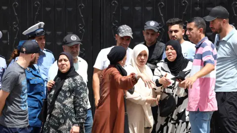 Police stand guard at a black gates of an orphanage in Algiers as a crowd of men and women gesticulate outside