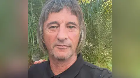 Ian Woolfenden wears a black polo shirt. He has short grey stubble and chin-length grey/brown hair with a short fringe. He is standing outside.