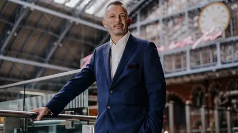 Transport UK Neil Grabham standing up in a railway station wearing a blue suit