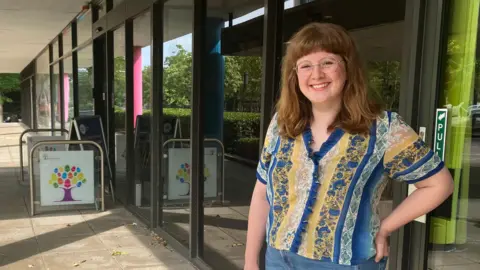 A picture of a woman with light brown shoulder length hair, who is wearing a colourful yellow and blue browse with flowers on it. She is wearing blue jeans and is stood outside Acorn House in Milton Keynes.