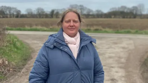 Dr Claire Brenner stands in front of an empty field wearing a blue coat and pink scarf. She has tied back blonde hair.