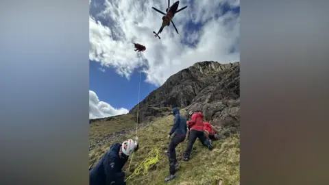 Keswick Mountain Rescue Team The casualty being winched up to the helicopter hovering above the slope. A man on the ground wearing a helmet is holding some yellow rope attached to the stretcher which is in the air. Three other mountain rescuers in red stand to the right.