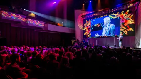 Wildscreen Sir David Attenborough is standing on stage at Wildscreen Festival, in front of an audience. He is speaking and gesturing with his hands and an image of him is also being projected on the screen behind him.