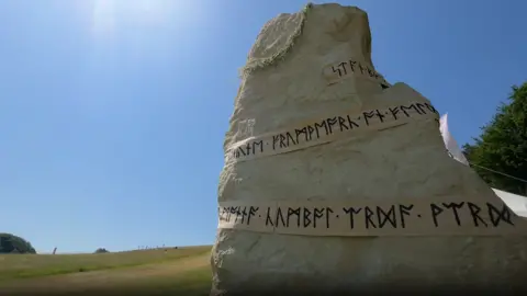 A large, beige-white sarsen stone in the landscape with blue sky behind it.