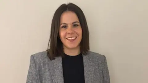 Silvia lleva a una mujer con cabello castaño con una chaqueta gris y una parte superior negra sonríe frente a una pared simple.