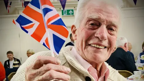 Matt Masters Dave Masters in a cream jumper smiling and waving a mini union flag on a stick. Behind him, there is flag bunting and a large flag with a crown on it. 