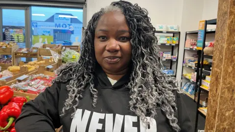 Kelly Foran/BBC Beatrice Zulu, who has black and grey curly hair and is wearing a black hoody with the slogan Never Give Up in white lettering, smiles at the camera inside a shop stocked with groceries.