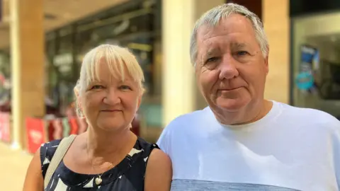 Shariqua Ahmed/BBC Amanda, a woman with short white hair, wearing a navy and white top, standing next to Frank, a man in a white T-shirt. They are looking into the camera and smiling.