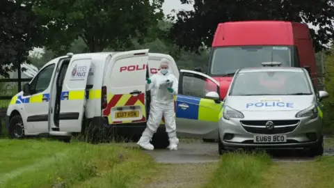 John Fairhall/BBC A forensic officer closing the door of a police van at the end of a grass driveway. A police car is parked next to it with open doors and a red van is in the background.