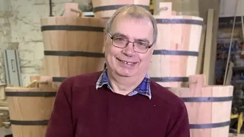 Allan Paulus wearing a red jumper and glasses. He's holding a bucket and is stood in front of a pile of buckets in his workshop