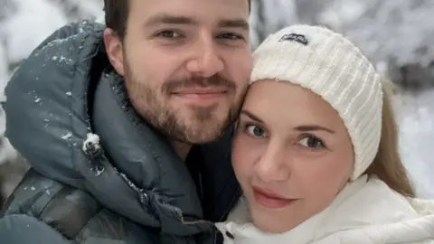 Two people standing close together in the foreground, wearing padded winter jackets, with one in a dark green coat and the other in a white coat and knitted headband. Snow is visible on the clothing. Behind them are snow-covered trees and branches.